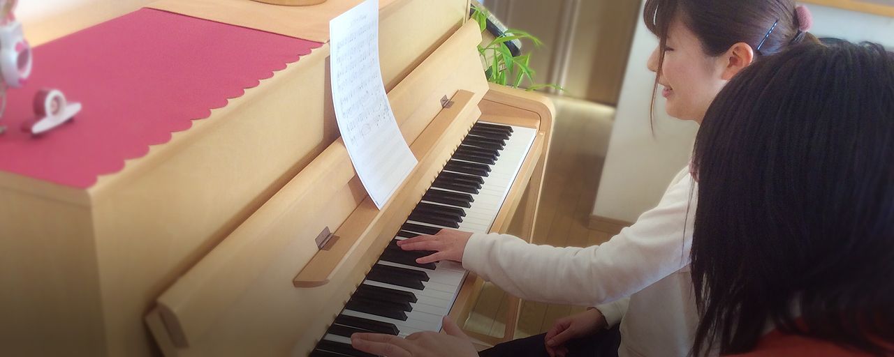 アイキャッチ画像：楽しそうに左手でピアノを弾いている若い保育士さんと指導している講師