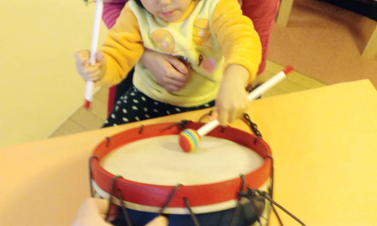 写真:お母さんの膝の上に座り、テーブルの上に置いてある太鼓をバチで叩く小さな子