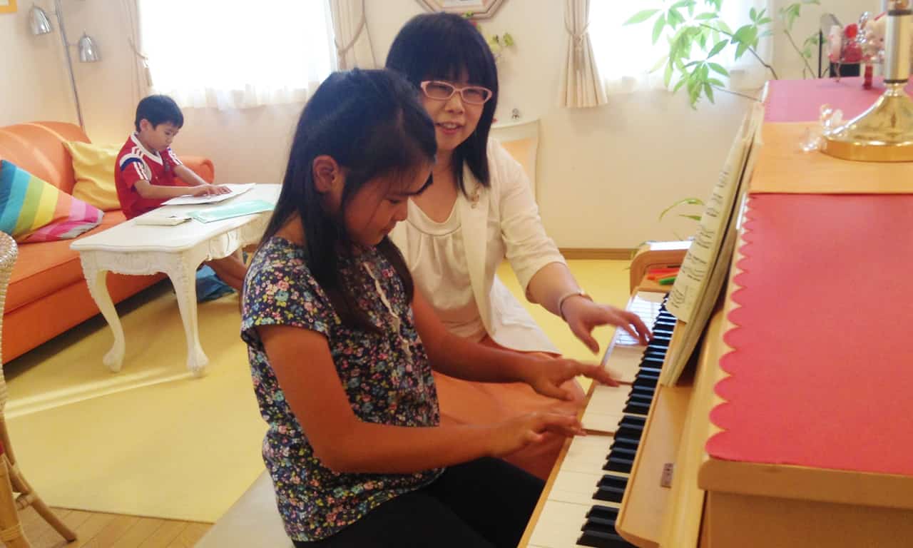 写真:両手でピアノを弾く女の子と、隣でサポートする講師
