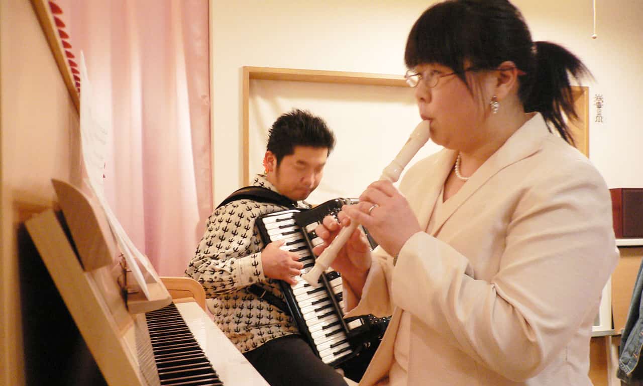 写真：アルトリコーダーを吹く当教室ピアノ講師の福田りえと、座ってアコーディオンを演奏している田ノ岡三郎さん