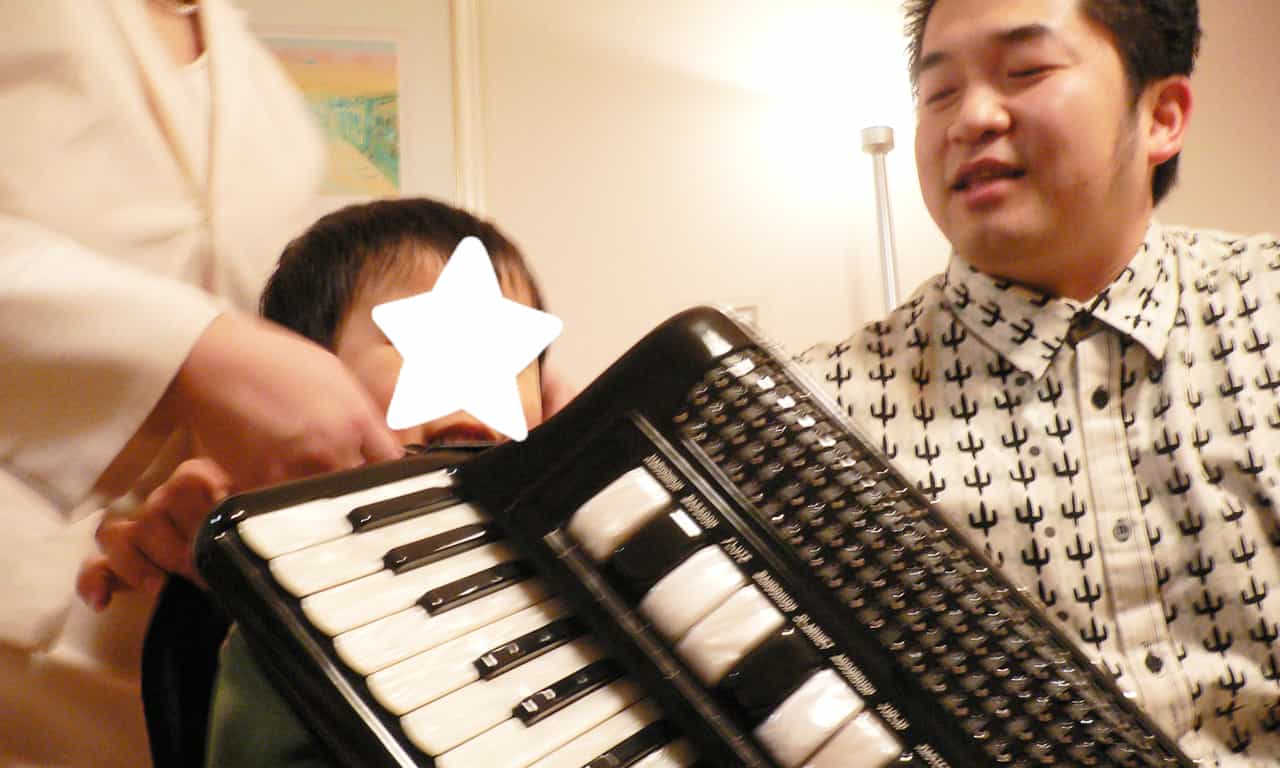 写真：大きなアコーディオンを肩に掛けようとする小さな男の子と、それを笑顔でサポートしている田ノ岡三郎さん