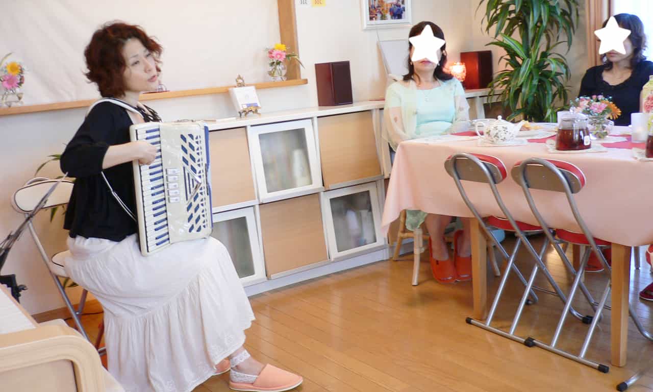 写真：歌いながらアコーディオンを弾くマルカートさんと、テーブル席で演奏に聴き入る大人の生徒さんたち