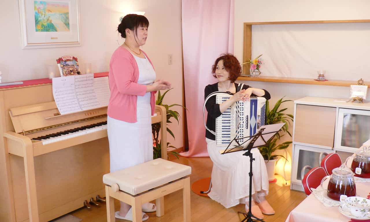 写真：椅子に座っているマルカートさんの側に立ち、マルカートさんの紹介をしている当教室ピアノ講師福田りえ