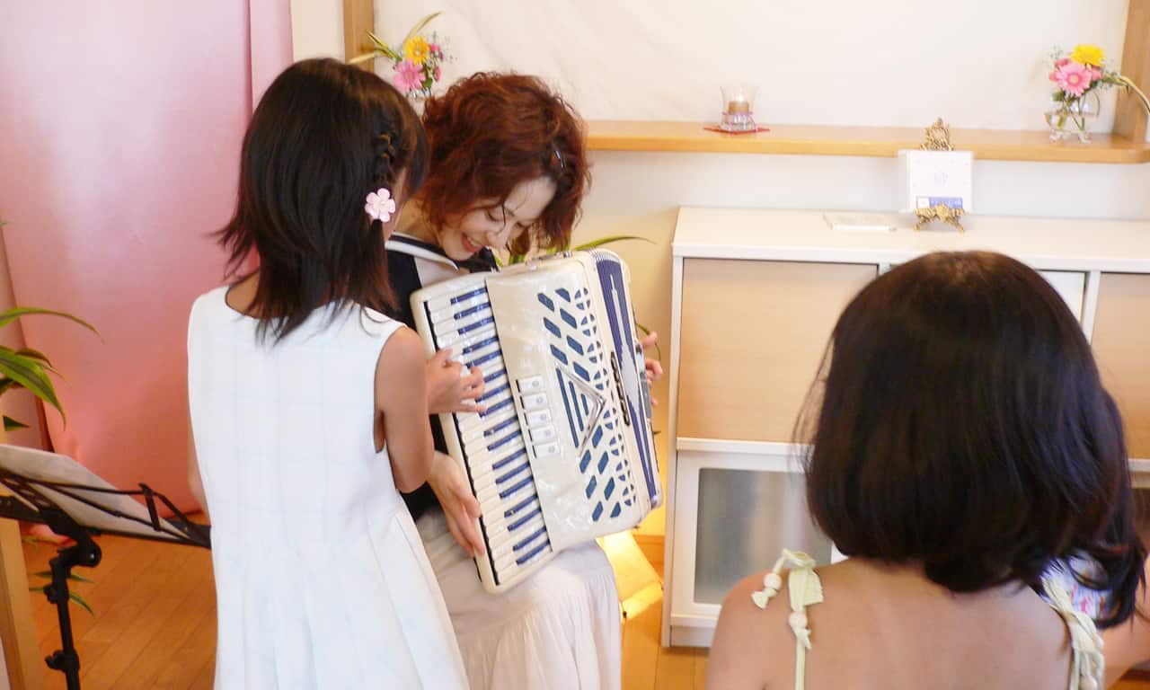 写真：笑顔のマルカートさんが支えているアコーディオンの鍵盤を、恐る恐る弾いてみる女の子と、後ろで順番を待っている女の子