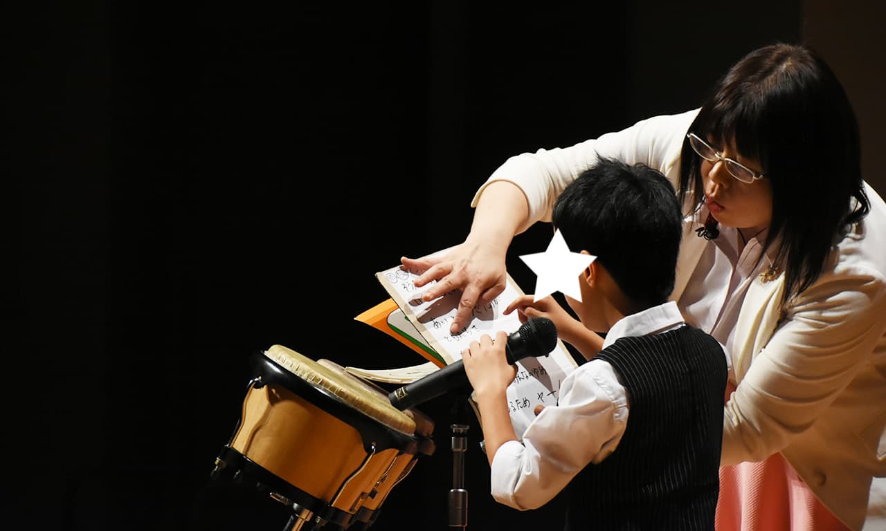 写真：マイクを握り、講師の開いた楽譜を指差し確認しながら歌っている男の子