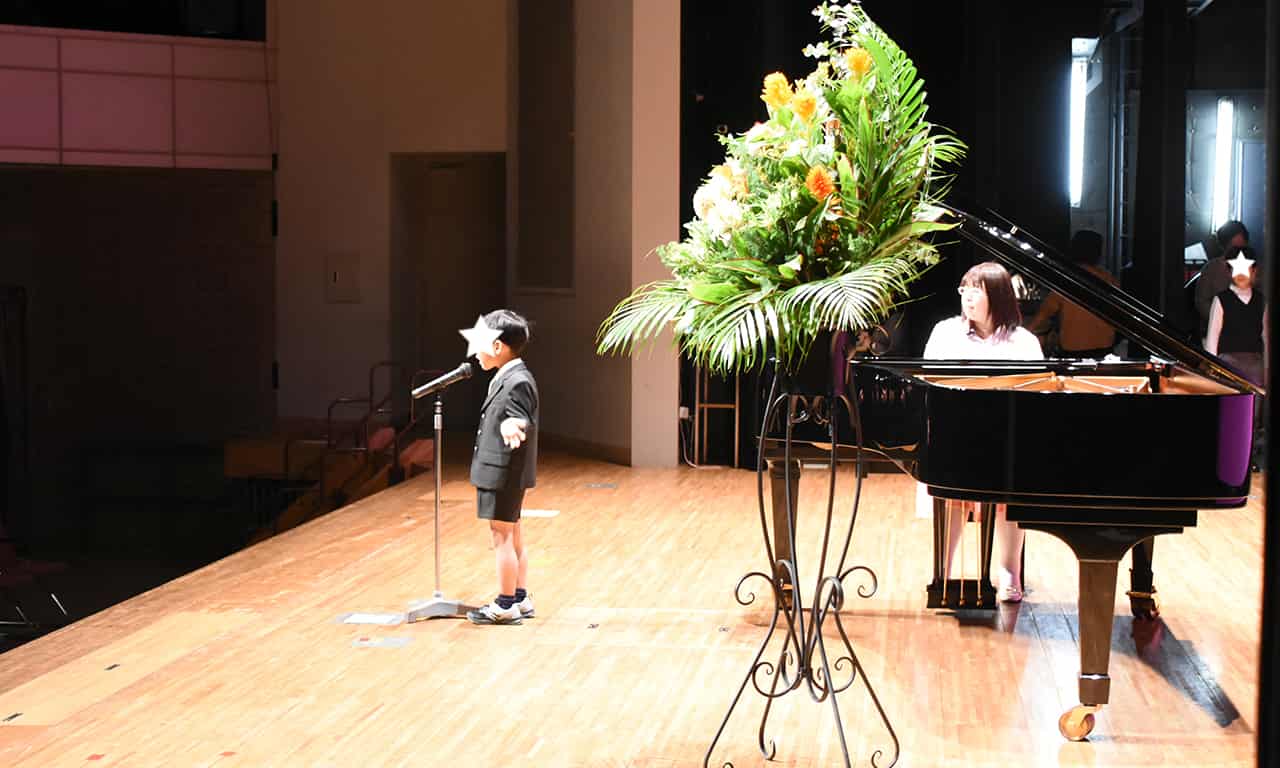 写真：スタンドマイクの前に立ち、両手を広げて歌うスーツ姿の男の子