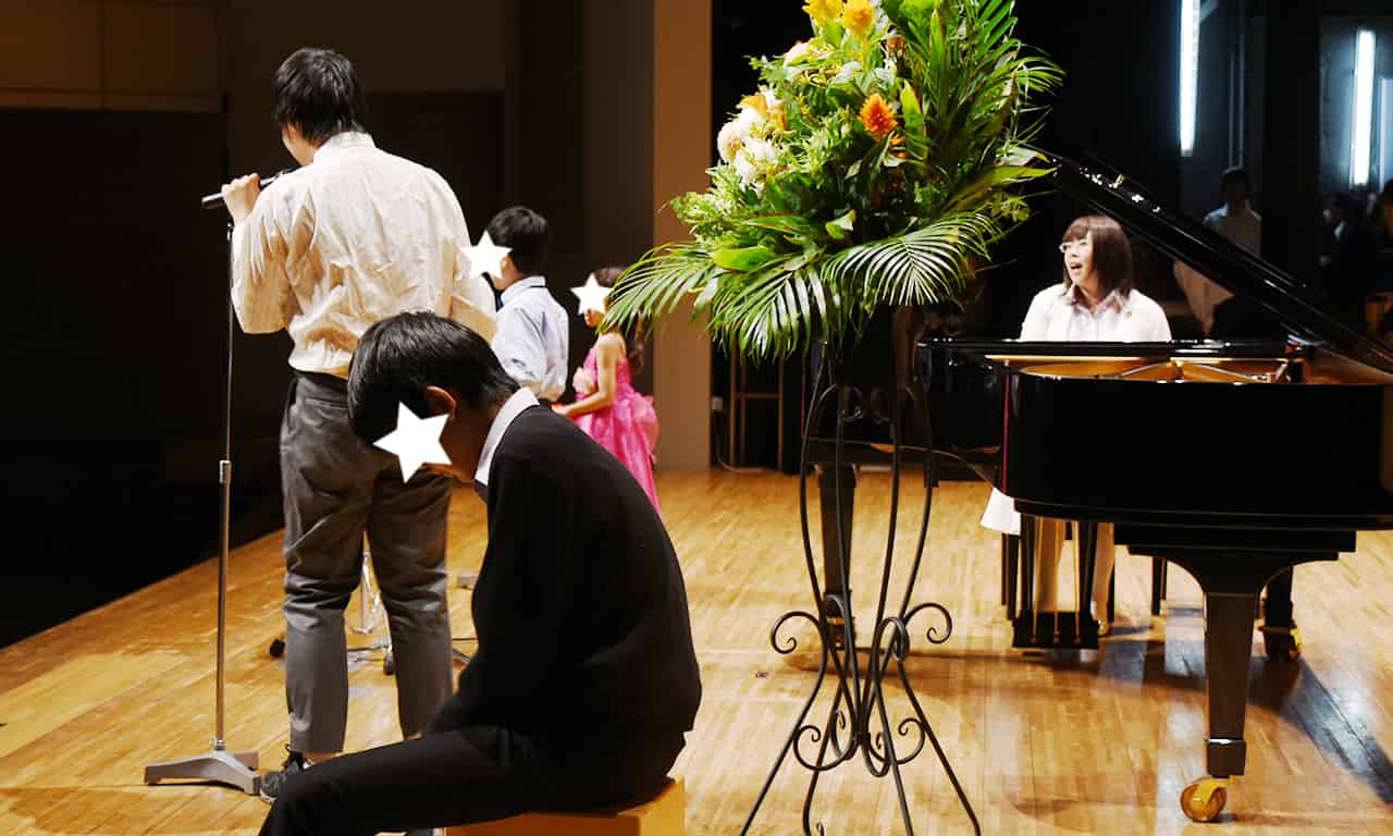 写真：ピンクのドレスを着た小さな女の子と、それよりすこしだけ大きな男の子、そして大きなお兄さんの三人がスタンドマイクで歌い、手前で男の子がカホンを叩き、講師がピアノを弾いている様子