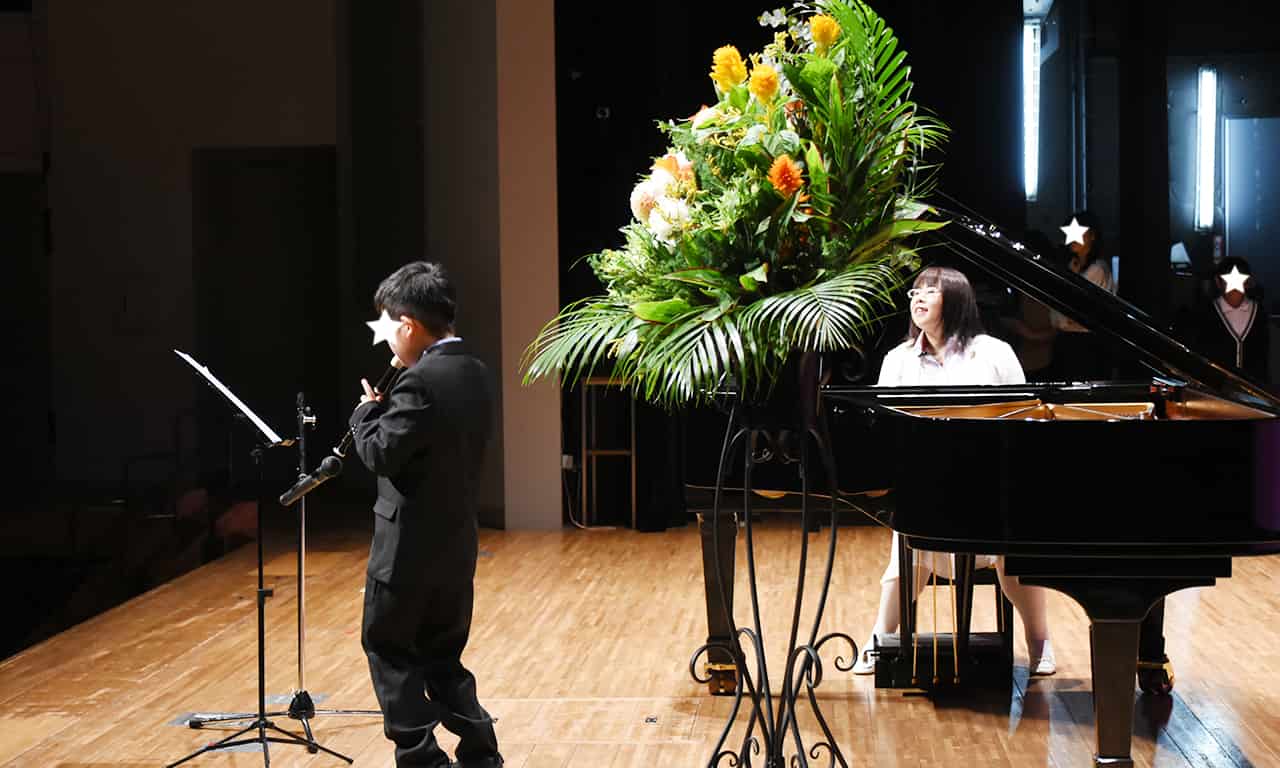 写真：一生懸命にリコーダーを吹いているスーツ姿の男の子と、笑顔で男の子を見ながらピアノ伴奏している講師