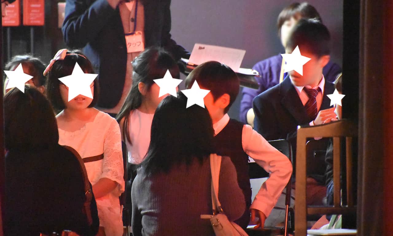 写真：舞台袖でイスに座って出番を待っている子どもたちの前で、かがみ込んで世話をしているお母さんたち