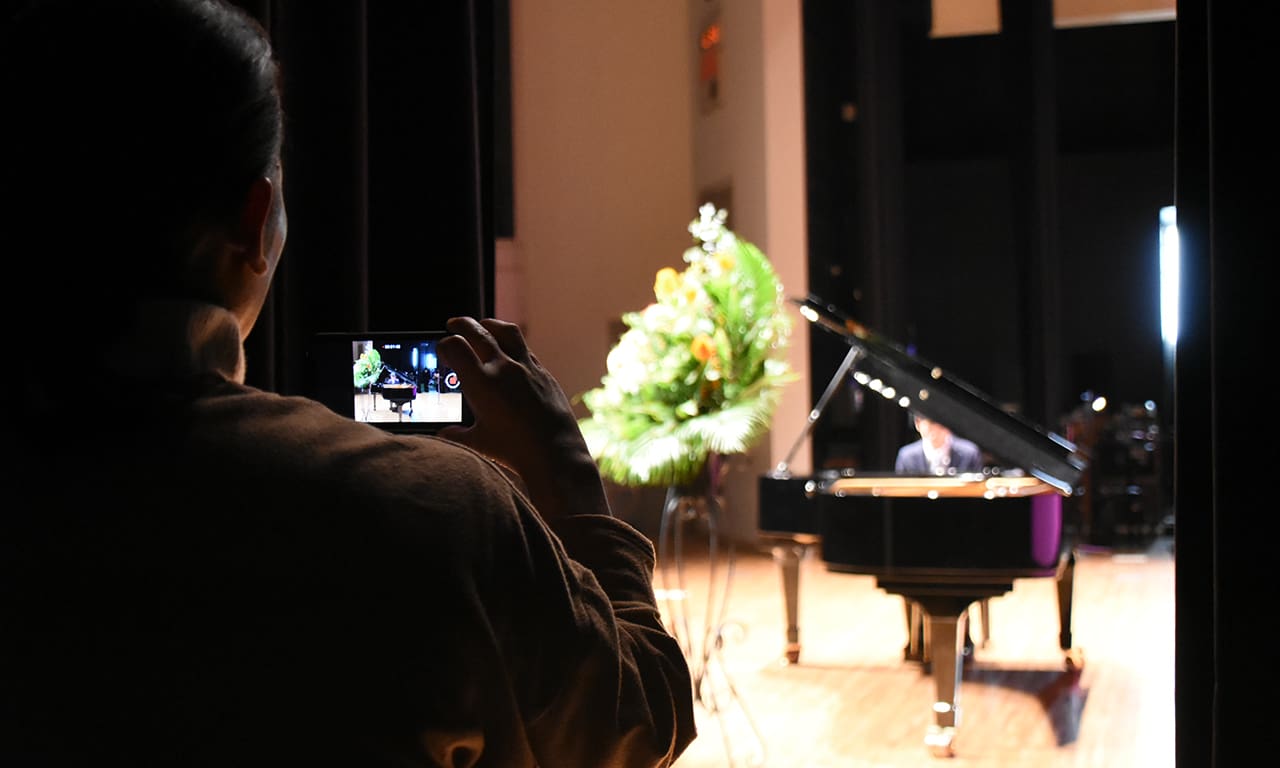 写真：ピアノ・ソロで演奏している男の子を、舞台袖から動画で撮影しているお母様の後ろ姿