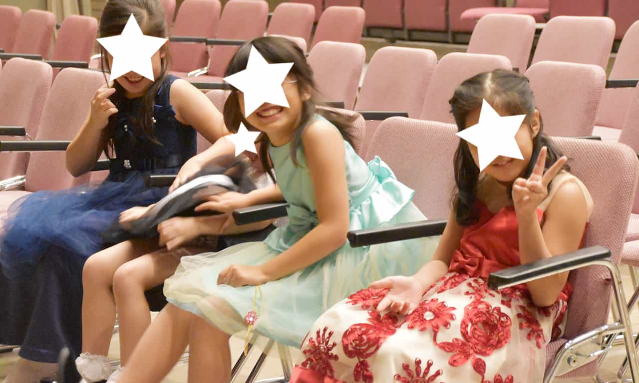 写真：会場のイスに座り、カメラにピースサインをしている、笑顔がはじける4人の女の子