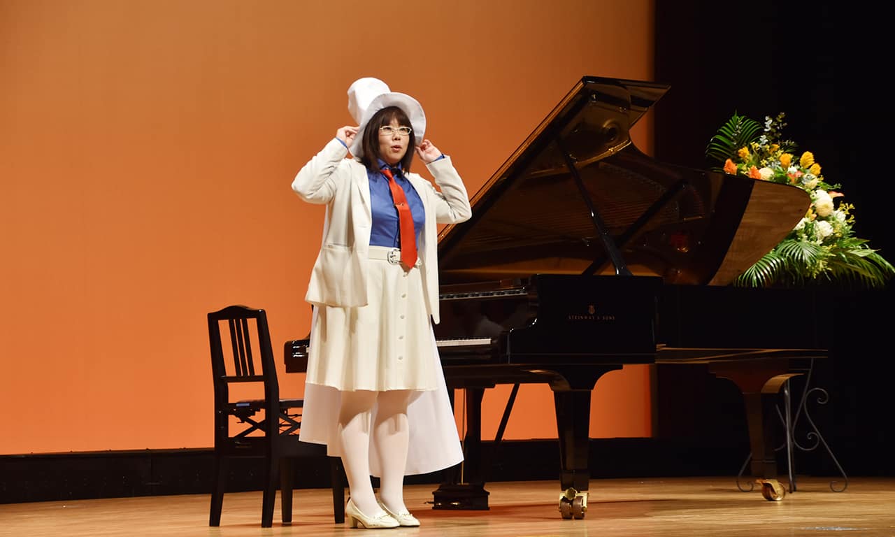 写真：白いシルクハットに白マント、白ジャケットと白スカート、青いシャツに赤いネクタイという衣装でピアノの前に立っている講師