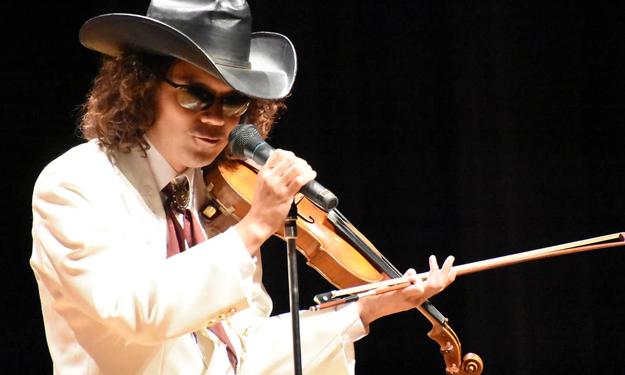 写真：ヴァイオリンを片手にマイクを握りトークを繰り広げている、白いジャケットに黒いテンガロンハットという出で立ちの穴澤雄介さん