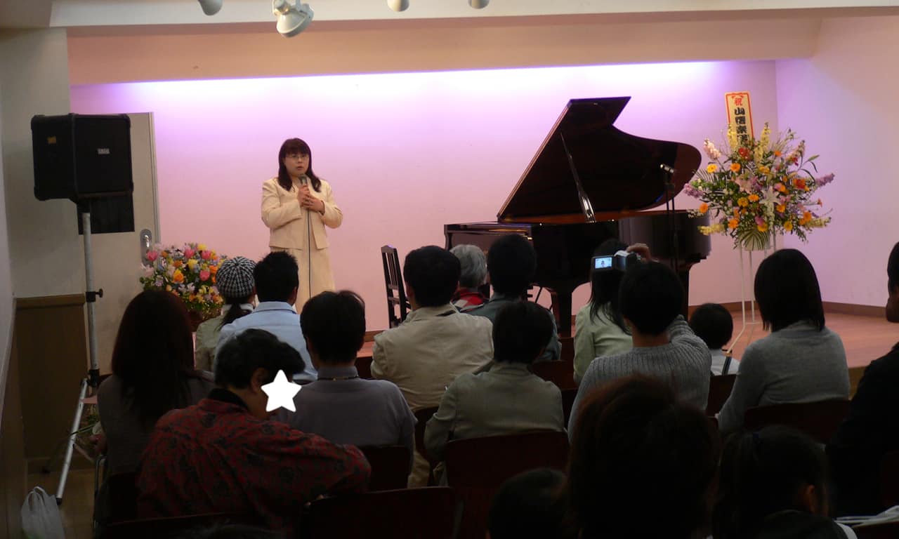 写真：マイクを持ってステージに立ち、来場者の方々に向かって発表会の挨拶を行っている講師