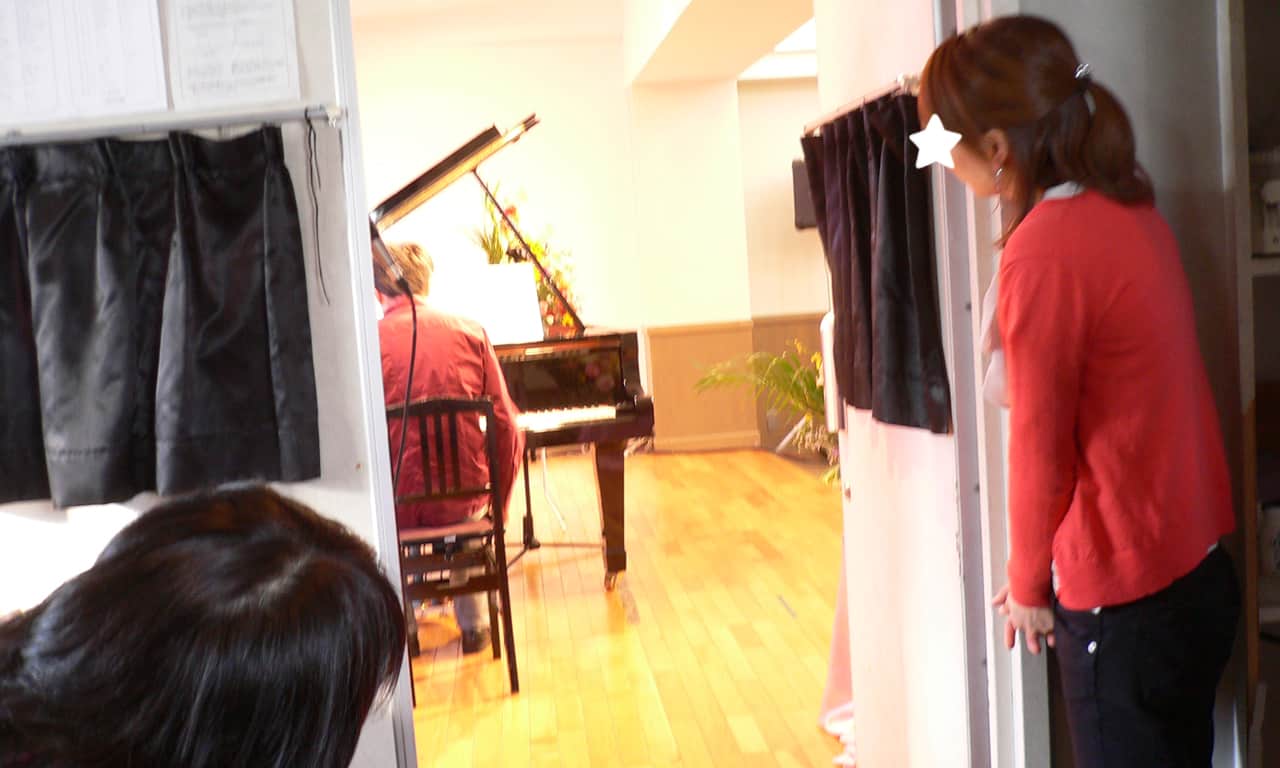 写真：演奏する大人の男性の後ろ姿を、舞台袖で見守る女性スタッフ