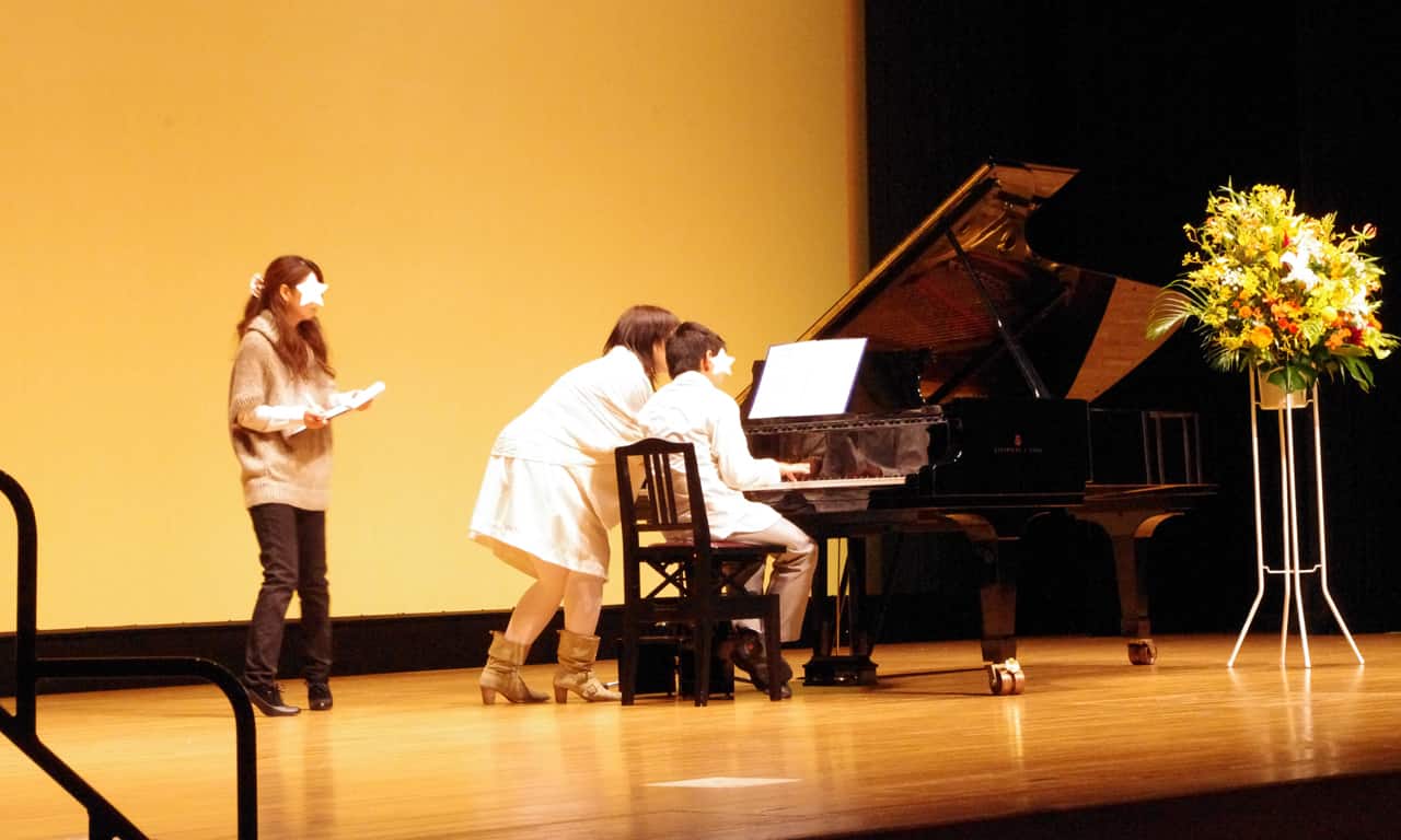 写真：ステージ上のグランドピアノでリハーサル演奏をする男の子と、指導する講師、見守るスタッフの様子