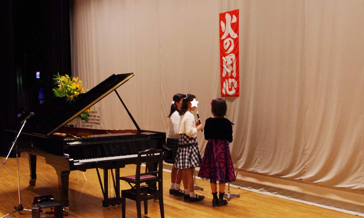 写真：緞帳が降りているステージで出番に備えて待機している、リコーダーを演奏する3人の女の子たち