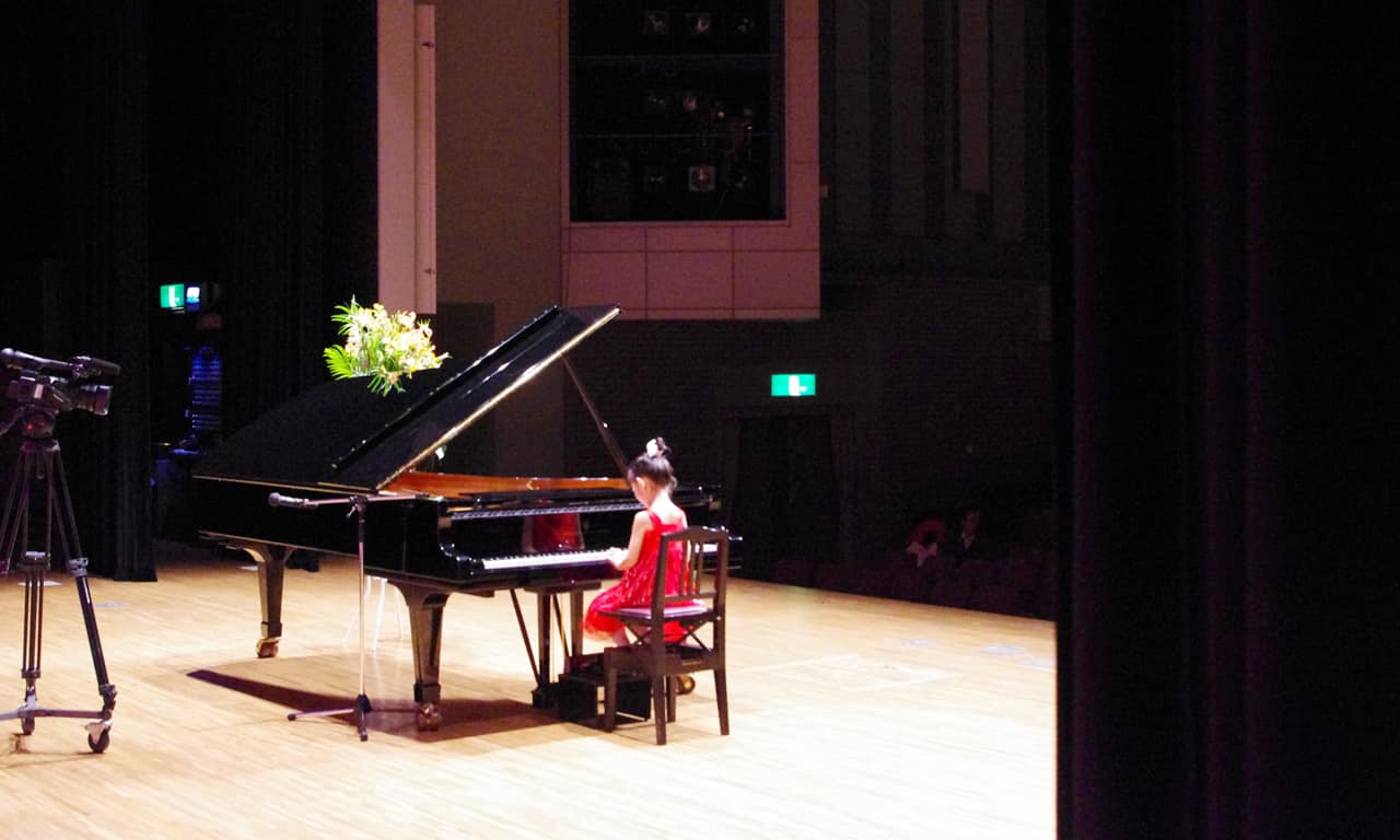 写真：広いステージで大きなグランドピアノ（スタインウェイ）を弾く、赤いドレスの小さな女の子
