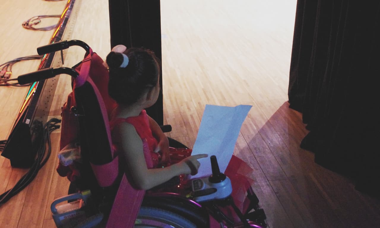 写真：舞台袖のカーテンの間からステージを見つめる車椅子の女の子