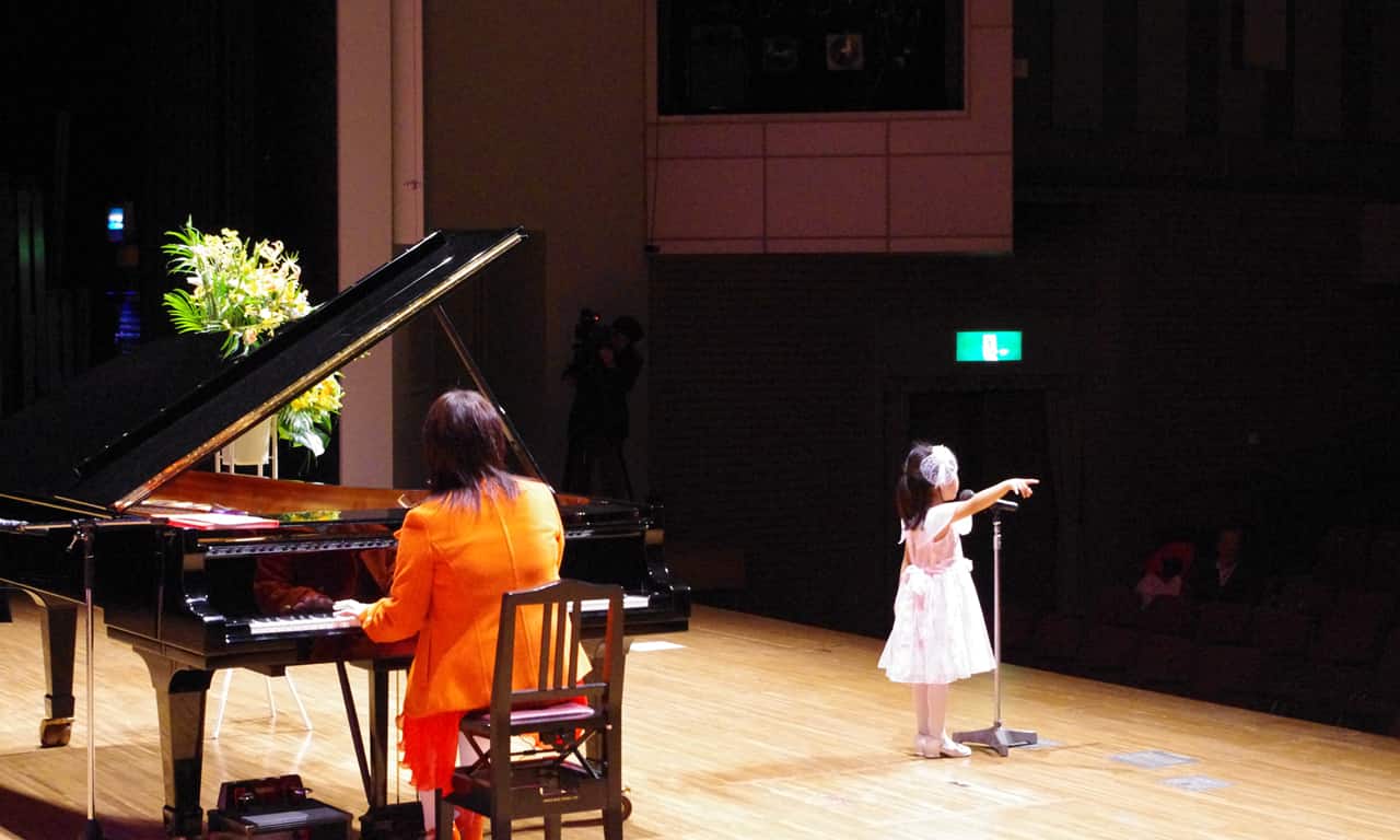 写真：講師のピアノ伴奏に合わせ、スタンドマイクの前で身振りを加えて歌う白いドレスの小さな女の子