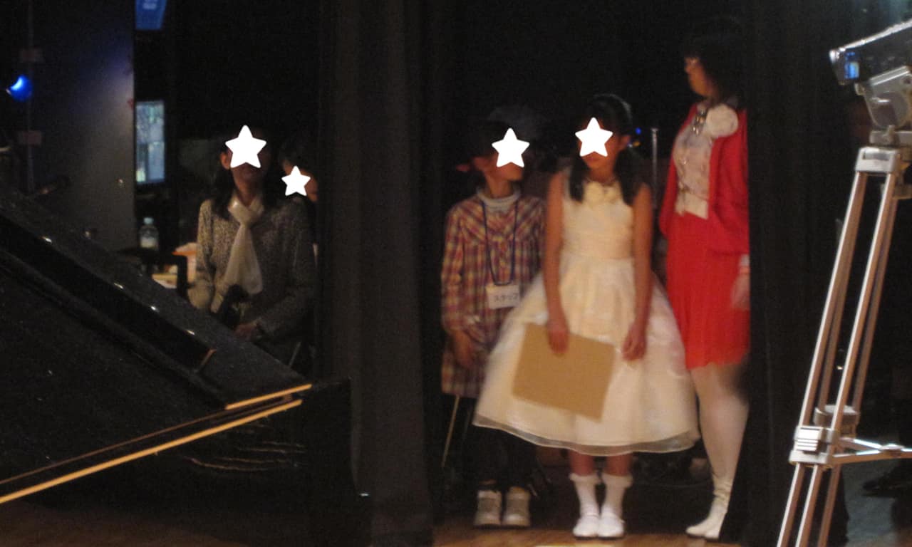 写真：舞台袖のカーテンの間で自分の出番を待っている、白いドレスの女の子とスタッフの様子