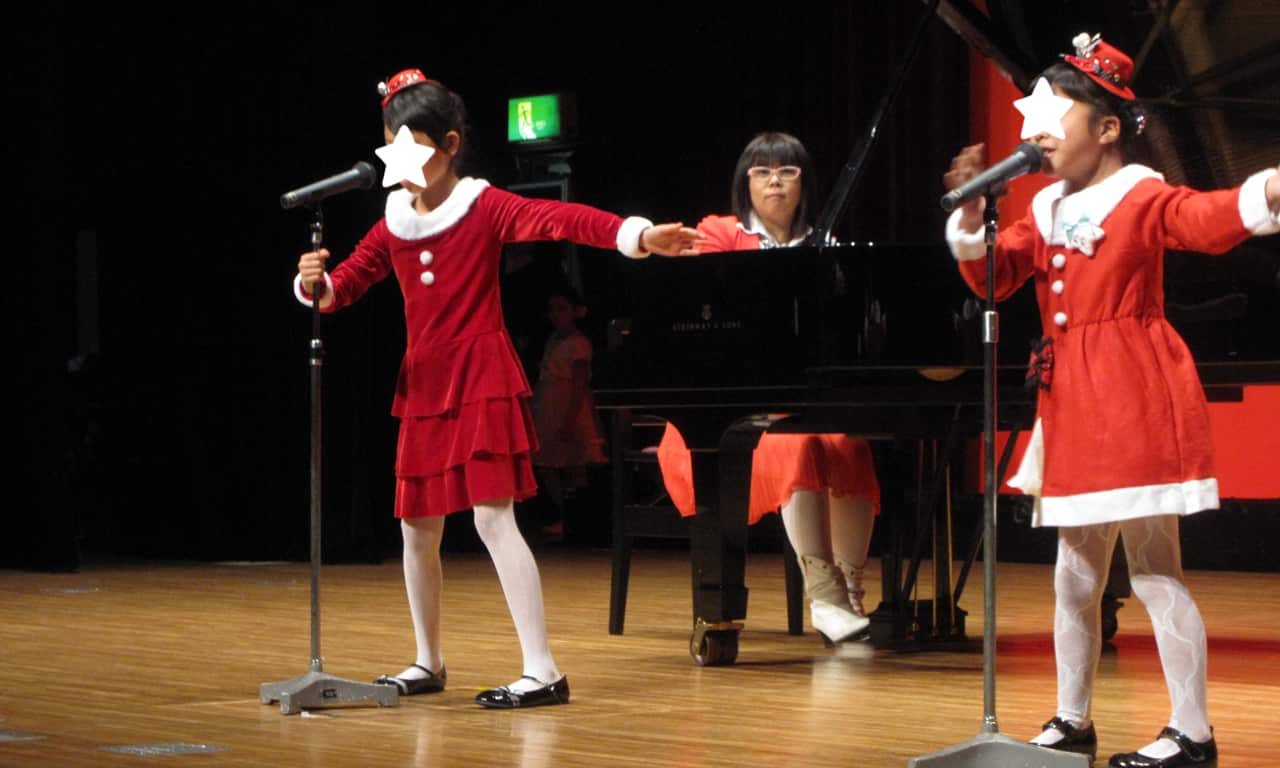 写真：スタンドマイクの前で振り付けと一緒に歌ってる、サンタクロース風の衣装の二人の女の子