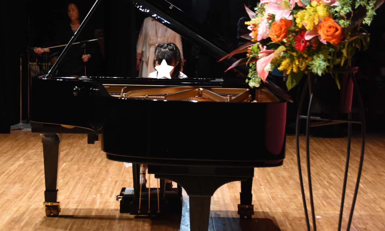 写真：グランドピアノ越しにピアノを弾く女の子