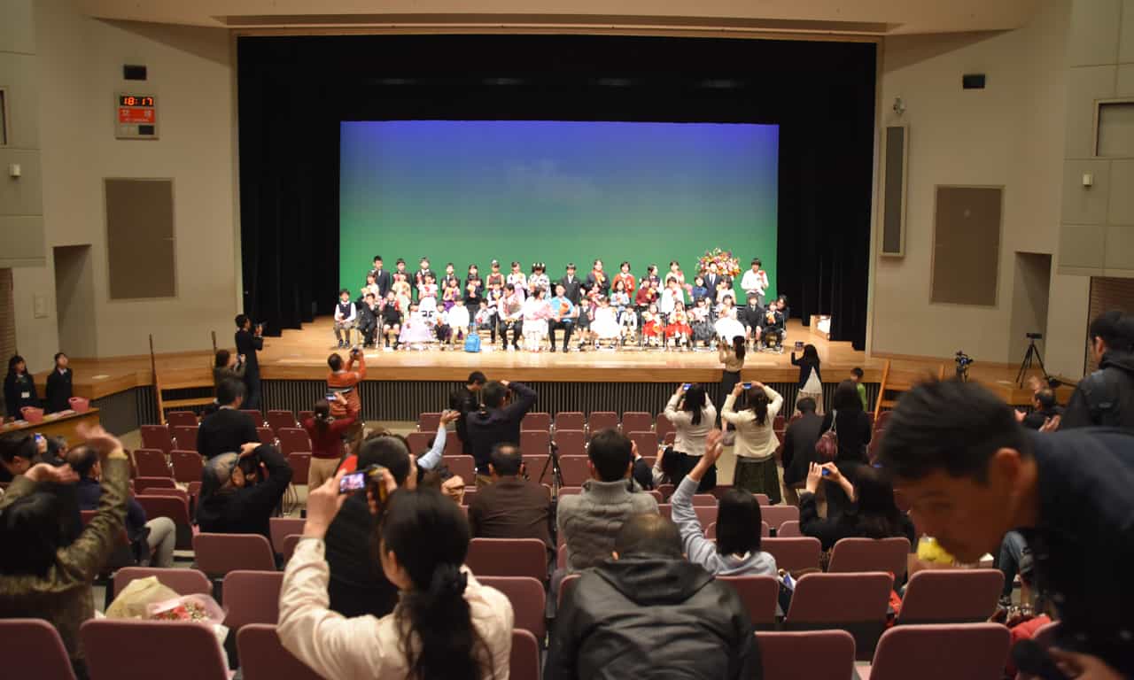 写真：客席最後方ろから見た会場の皆さんによる記念撮影の様子。多くの親御さんが手にスマホやデジカメを持ち、ステージ上に整列している出演した子どもたちを撮影しています。